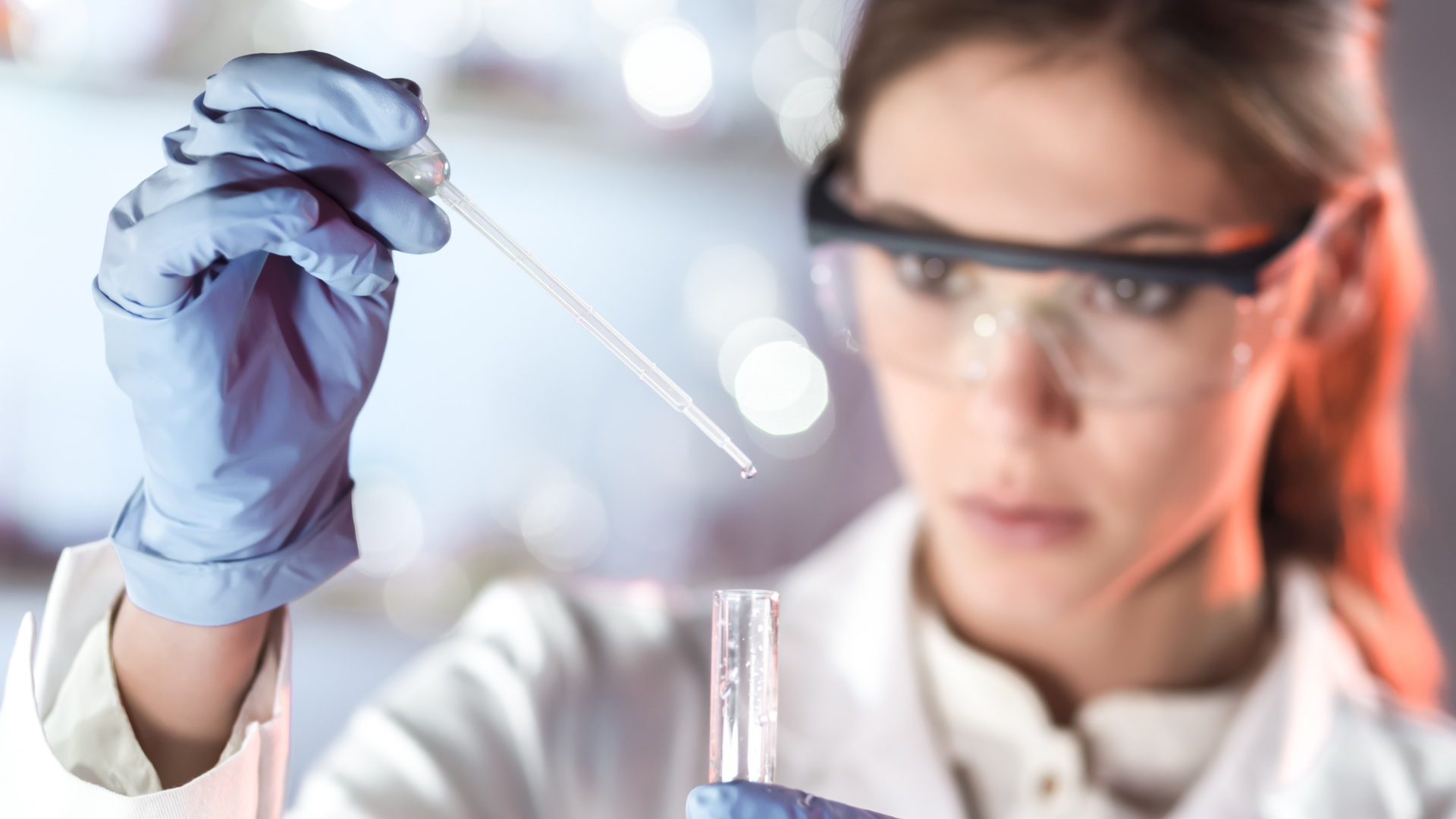 Young woman in lab coat and safety goggled holds a dropper